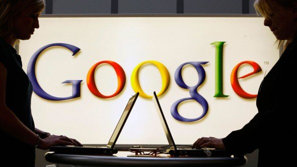 Silhouettes of two individuals working on laptops in front of a brightly lit Google logo.