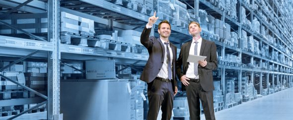 Two men in suits discussing logistics in a warehouse with shelves filled with boxes.