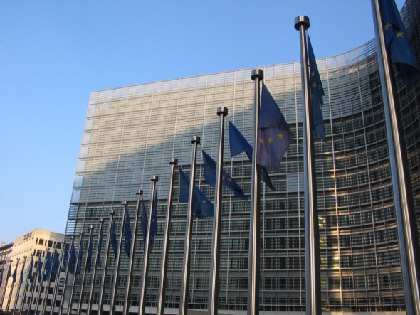 A modern glass building of the European Commission with several flags of the European Union displayed in front.