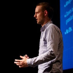 A man in a checkered shirt speaking on stage with a blue presentation in the background.