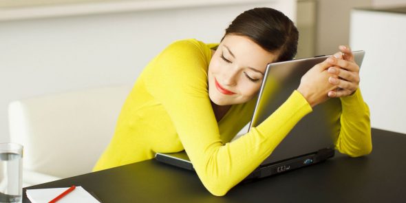 A woman in a bright yellow sweater smiling while hugging a laptop on a table.