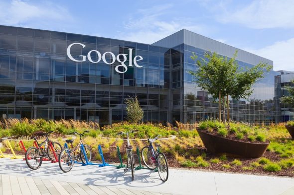 A view of the Google campus with bicycles parked in front of a modern glass building featuring the Google logo.