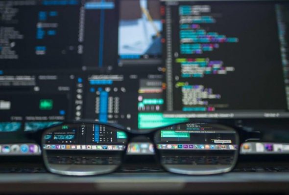 A close-up view of glasses reflecting lines of code and data analysis on a computer screen.