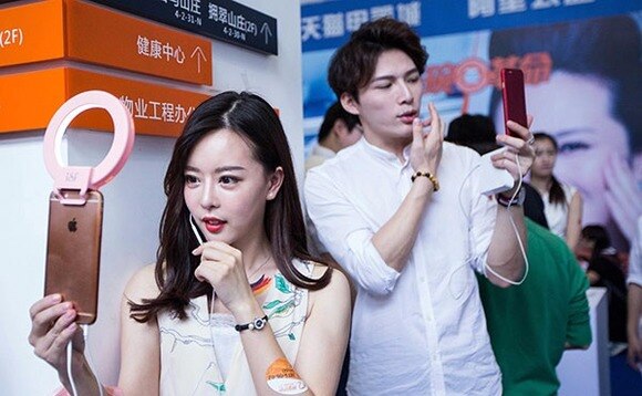 Two individuals at an event taking selfies with phones; the woman has a ring light attached to her phone.