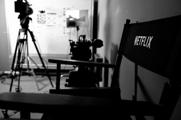 A black and white image of a director's chair with 'NETFLIX' printed on the back,positioned in front of film equipment.