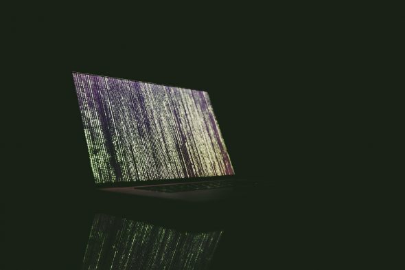 A low-light image of a laptop displaying matrix-like code on its screen,reflecting on a surface.