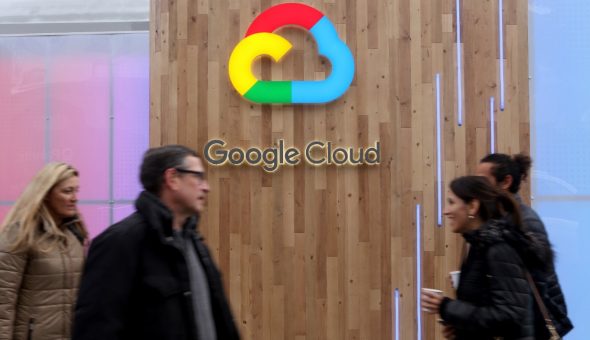 A sign of Google Cloud displayed on a wooden wall with people walking in front.