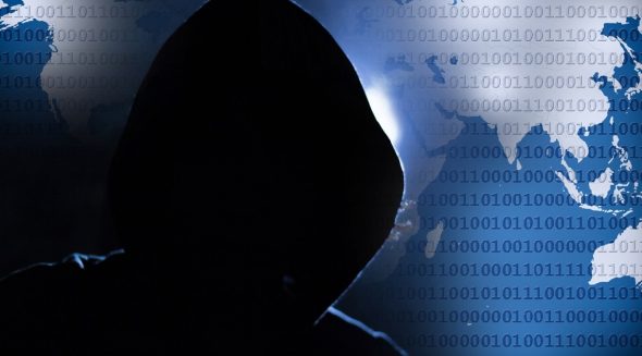 A silhouette of a figure wearing a hood in front of a world map,overlaid with binary code,symbolizing cybercrime or hacking.