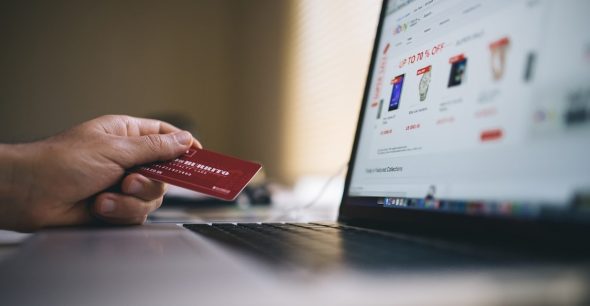 A hand holding a credit card in front of a laptop displaying an online shopping website.