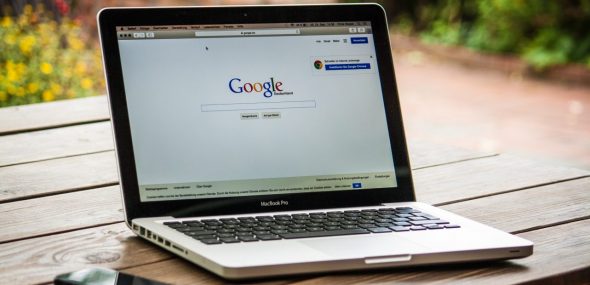 A MacBook Pro displaying the Google search page,placed on a wooden table outdoors.