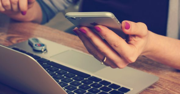 A person's hand holding a smartphone while using a laptop