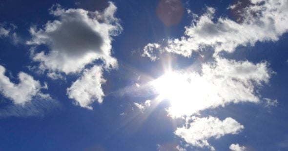 A bright sun shining through fluffy clouds against a clear blue sky.