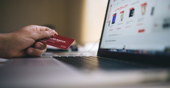 A hand holding a credit card in front of a laptop displaying an online shopping website.