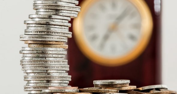 A stack of coins arranged neatly in multiple rows with a blurred clock in the background.