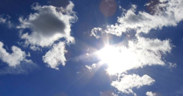 A bright sun shining through fluffy clouds in a clear blue sky.