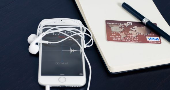 A white smartphone with tangled earphones,a credit card from Deutsche Bank,a notebook,and a pen arranged on a dark surface.