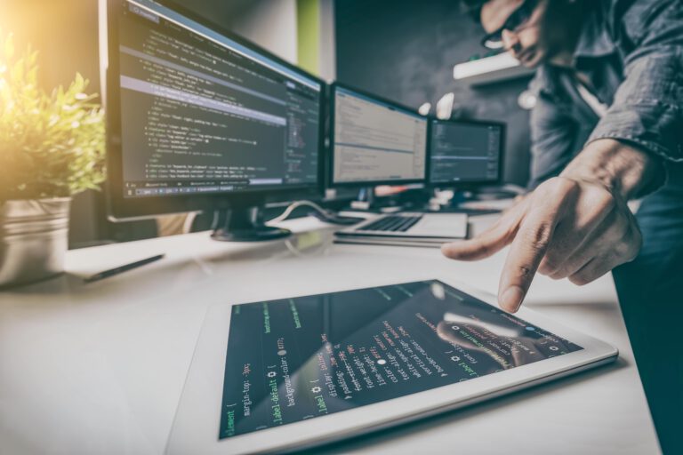 A person pointing at code on a tablet with multiple monitors in a tech workspace.