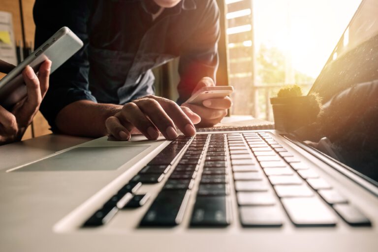 Close-up of hands using a laptop and smartphones in a well-lit workspace.