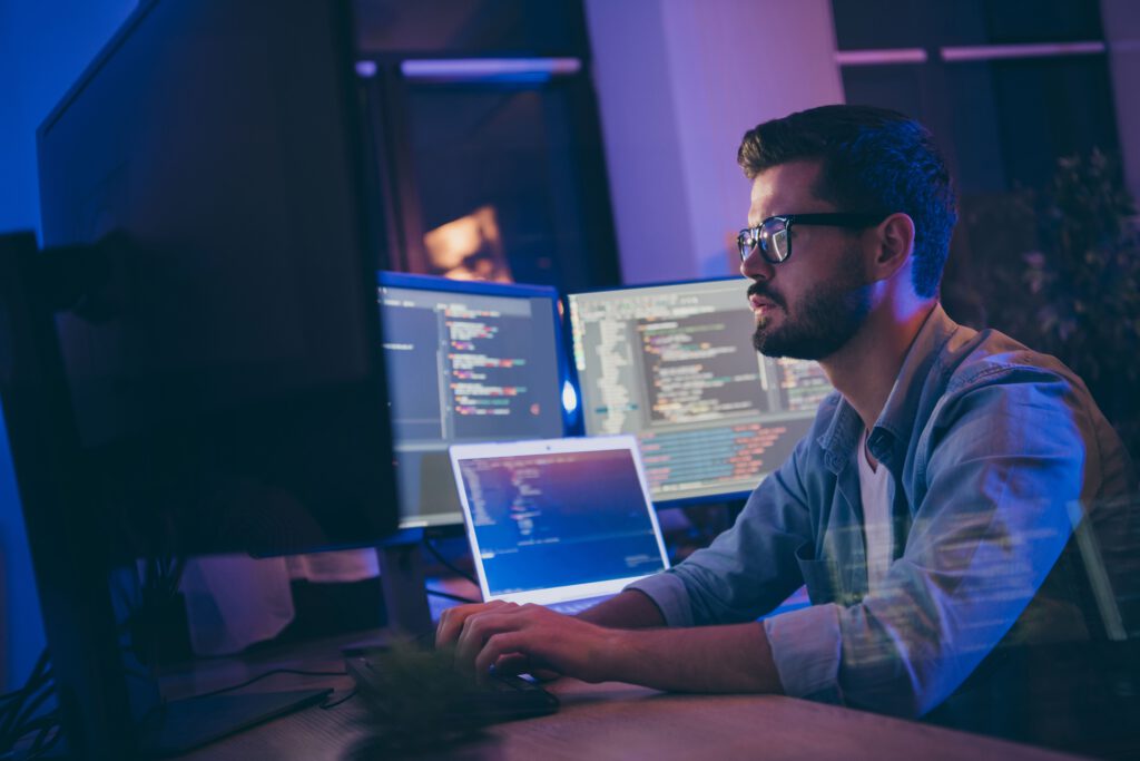 A focused programmer working on a laptop in front of multiple monitors displaying code,illuminated by a blue light.