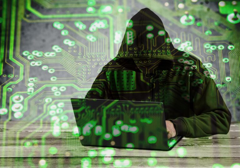 A hooded figure working on a laptop with digital circuit patterns overlaying the image.