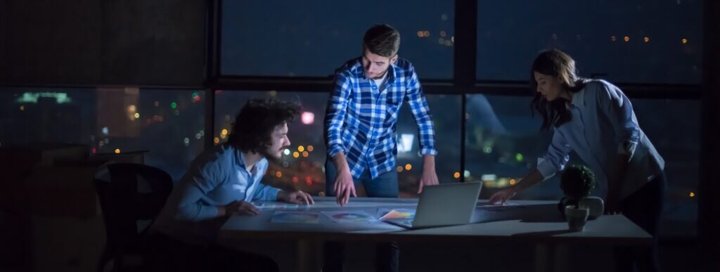 Three people collaborating over documents on a table in a dimly lit room with a city view at night.