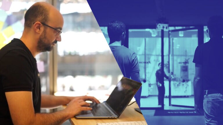 A man typing on a laptop, with people entering through doors in the background.