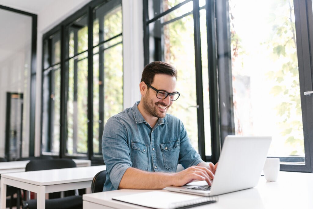 Smiling man in casual attire using a laptop at a bright workspace.