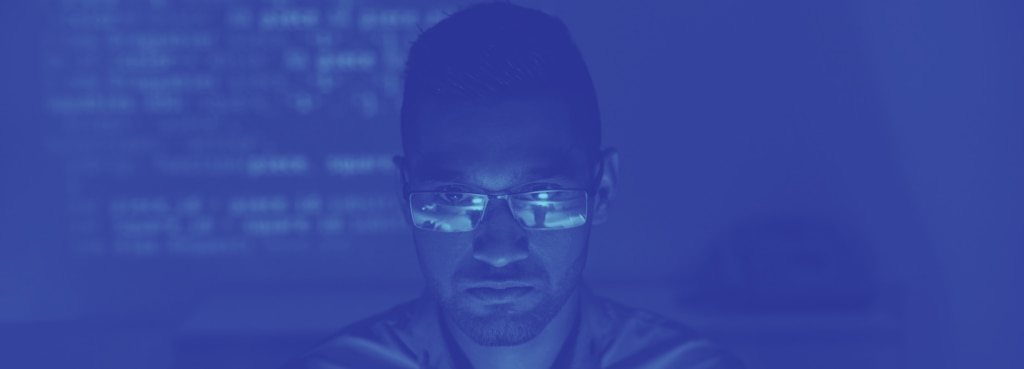 A person wearing glasses focused on a computer screen illuminated by a blue light.