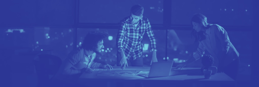 Three individuals collaborating over a table with a laptop and a cityscape view at night.