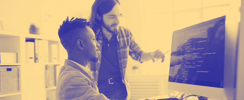 A man is giving instructions to another individual at a computer,presumably demonstrating coding or programming.