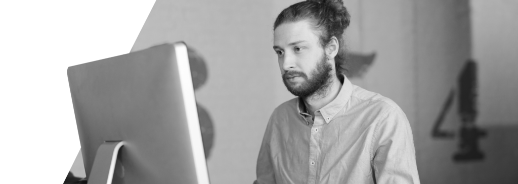 A focused man with a beard and long hair in a bun,sitting at a desk working on a computer.