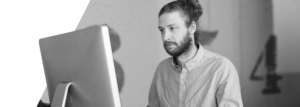 A focused man with a beard and long hair in a bun,sitting at a desk working on a computer.