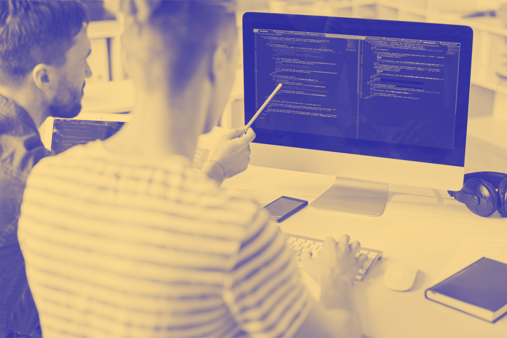 Two individuals collaborating on coding at a computer,one pointing at the screen with a pen.