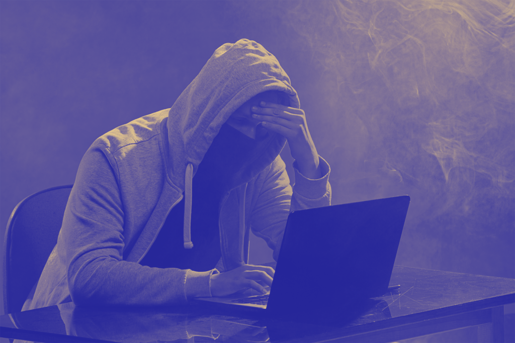 A person in a hoodie is sitting at a desk with a laptop,appearing stressed or frustrated,set in a moody atmosphere.