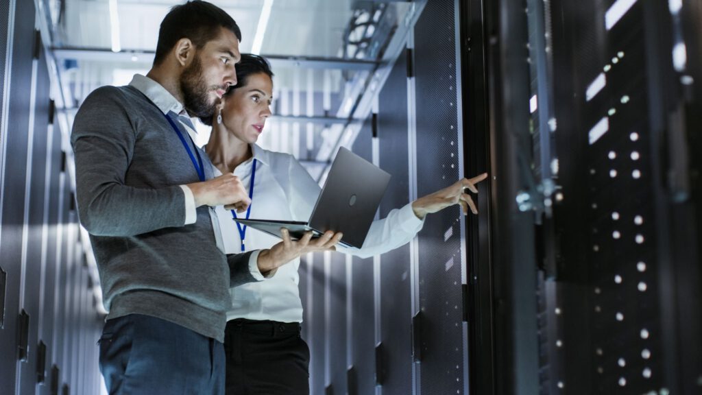 Two professionals collaborating in a data center,discussing tasks while using a laptop.