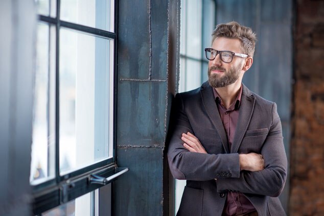 A bearded man in a suit and glasses,standing with his arms crossed while looking out of a large window.