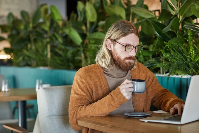 A man with long hair and a beard wearing glasses is sitting at a table in a cafe