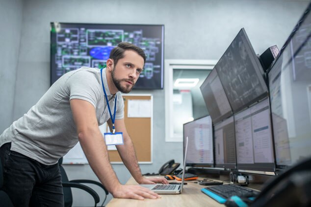 A focused man working on a laptop in a control room,surrounded by large monitors displaying various data and information.