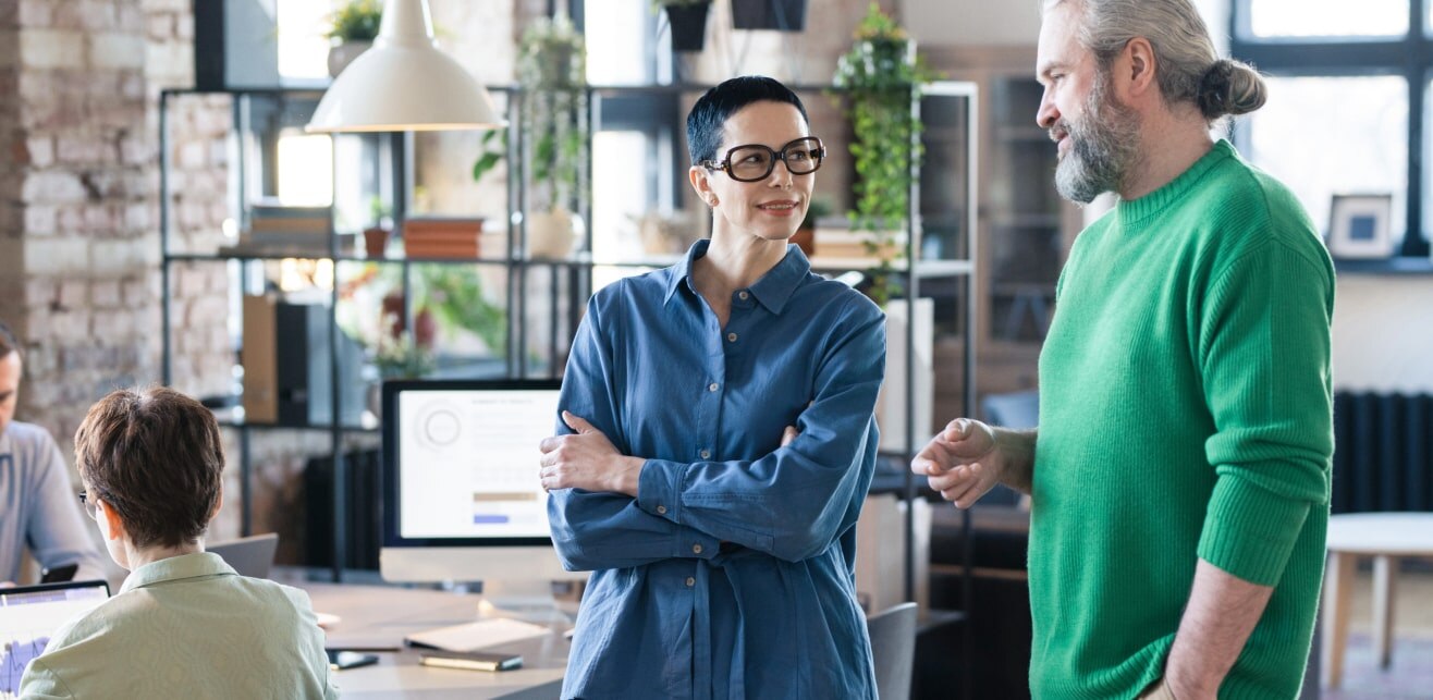 A professional setting with two individuals engaging in conversation while others work at desks in the background.