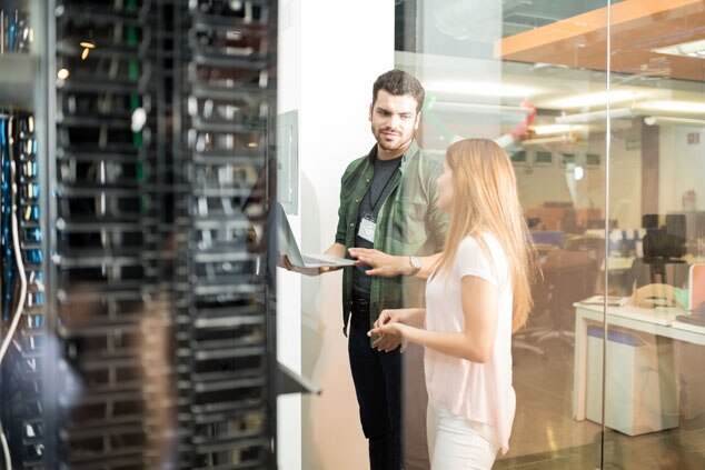 A man and a woman discussing in an office setting,next to a server room,while using a laptop.