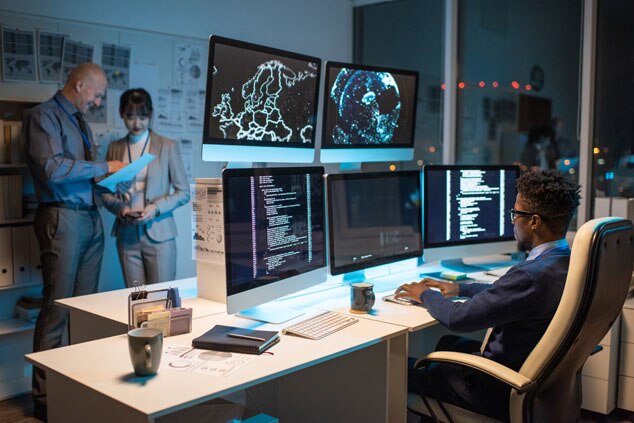 A modern office with multiple screens displaying data and two people discussing.