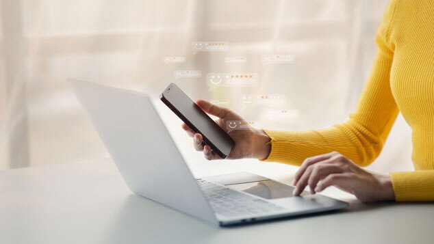 A person in a yellow sweater holds a smartphone while typing on a laptop