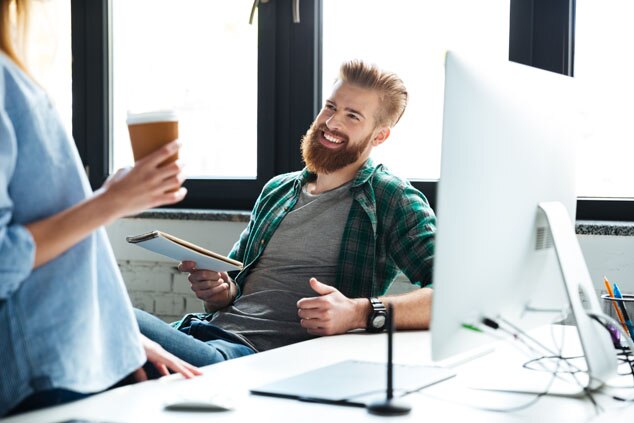 A smiling man with a beard sitting in an office,holding a notepad,while a woman with a coffee cup is standing nearby.