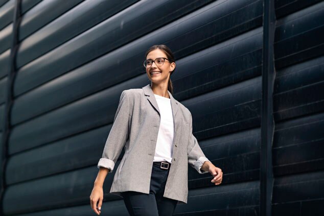 A woman in a gray blazer and white shirt,smiling while walking against a modern black wall.