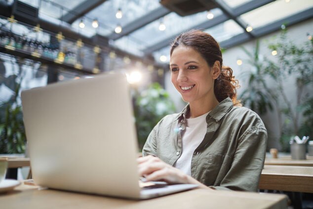 A woman smiling while using a laptop in a bright,green indoor setting.