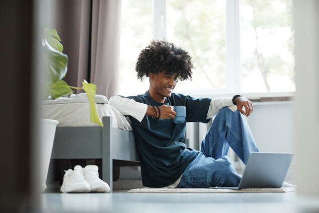 A young person sitting on the floor with a laptop,smiling while holding a cup,in a cozy room.