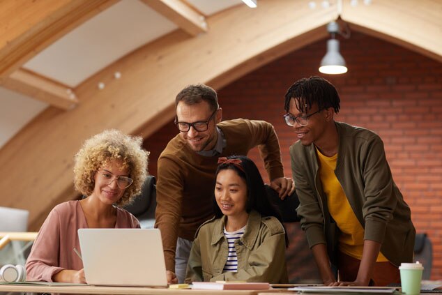 A diverse group of four people engaged in a collaborative discussion around a laptop in a modern workspace.