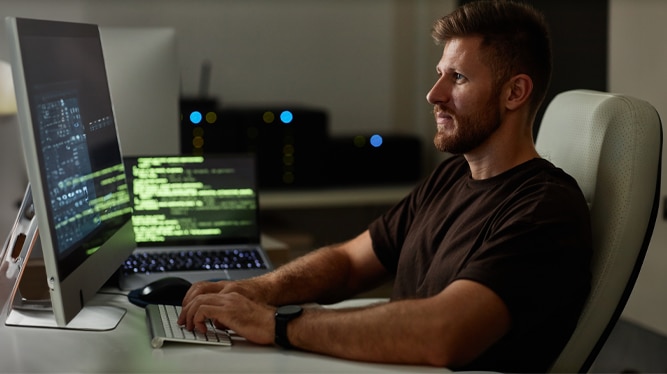 A man sitting at a desk,focused on coding on a computer screen in a dimly lit environment.