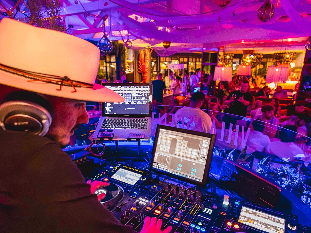 A DJ wearing a large hat mixes music at a vibrant nightclub with colorful lighting and a lively crowd in the background.