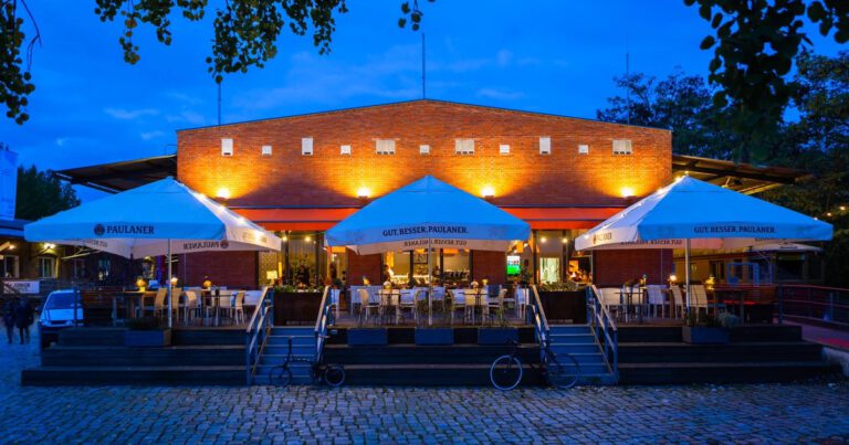 A modern brick restaurant with outdoor seating under large umbrellas,illuminated at dusk with warm lighting.